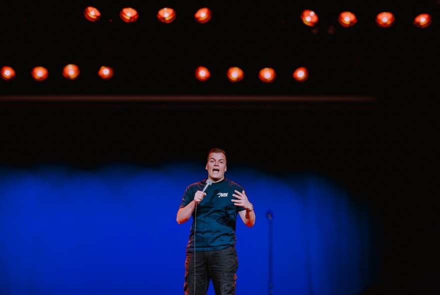 Comedian performing on stage under lights and in front of blue curtains, holding a microphone and gesticulating.