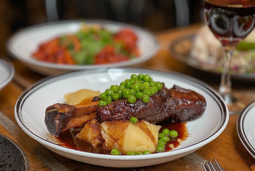 A meat, potatoes, peas and gravy dish on a wooden table with a glass of red wine next to it.