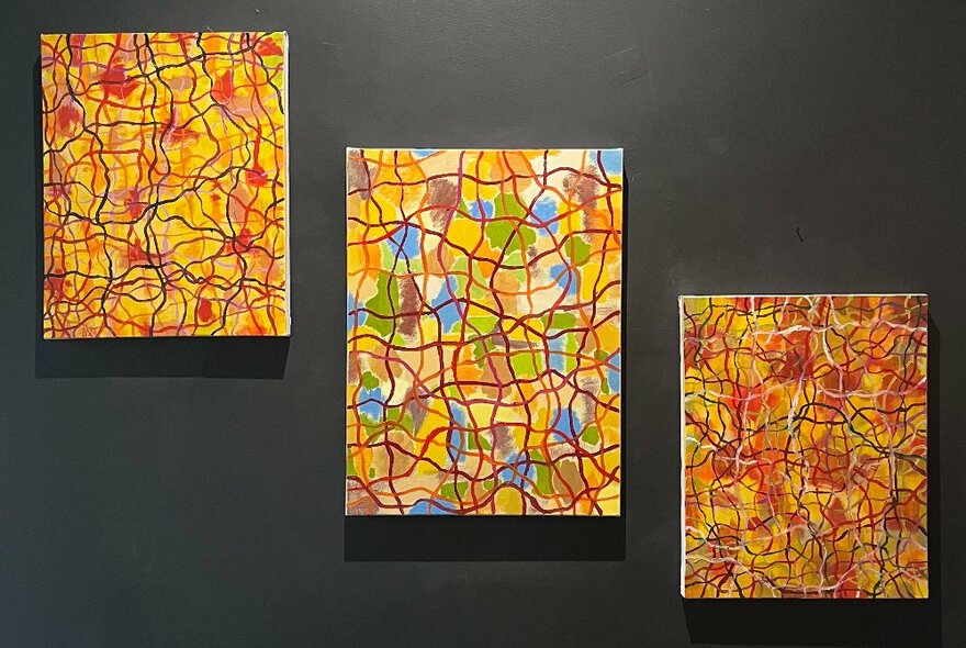 Three abstract paintings composed mostly of intersecting lines, in shades of yellow and red,  hanging on a dark wall in a gallery space.