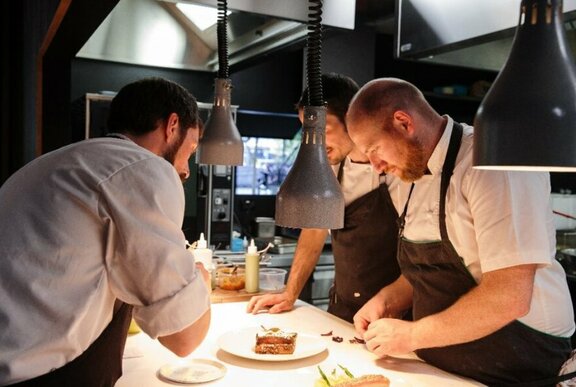 Three chefs working together around a task-lit workbench, adding the finishing touches to a culinary dish.