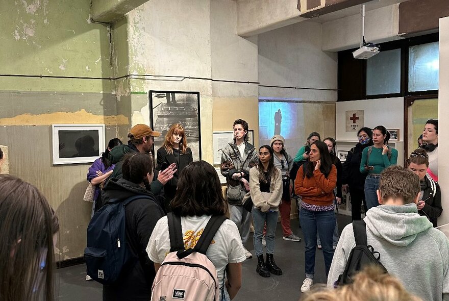 A group of people standing around in a room, listening to a person talking, with objects and artworks displayed on the walls.