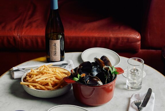 Marble restaurant table set with bowls of chips and mussels, with cutlery, napery and a bottle of wine.