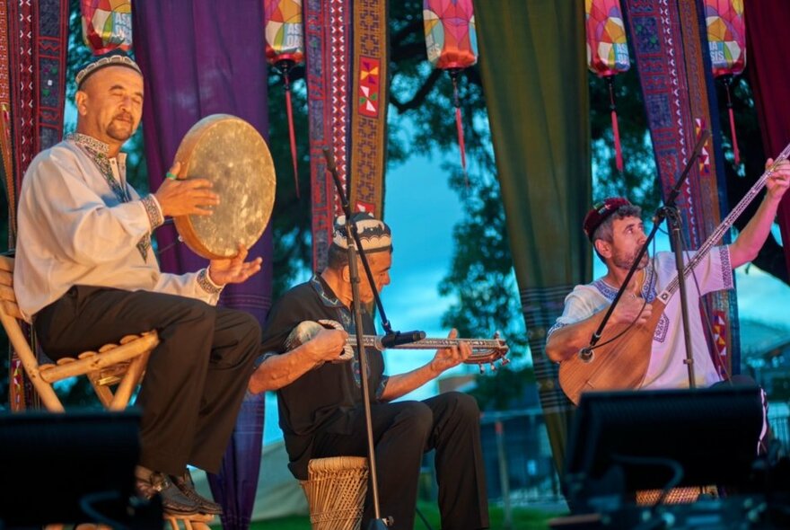 Three musicians playing instruments on an outdoor stage in a twilight setting.