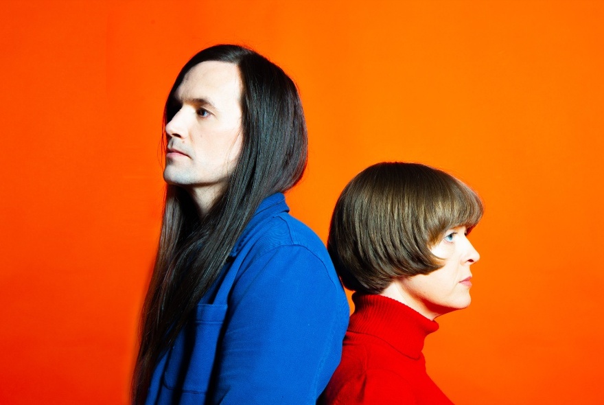 Two people standing back to back in profile, one person with long straight hair wearing a bright blue jacket, the other with a bob haircut wearing a red jumper, with an orange background behing them.