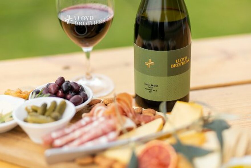 An antipasto platter, a bottle of red wine and a glass of red wine all resting on a wooden table.