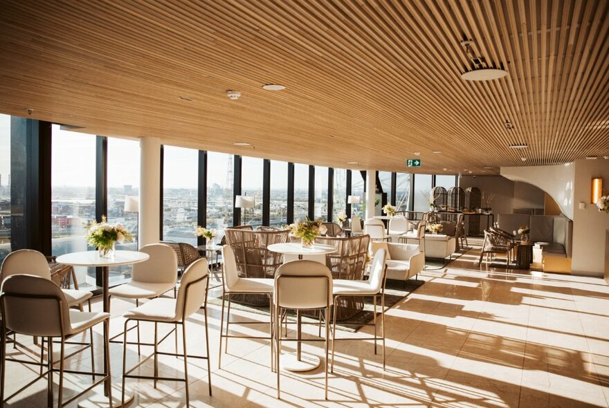 Sun-drenched penthouse lounge bar with tables and chairs, panoramic windows and striped wooden ceiling.