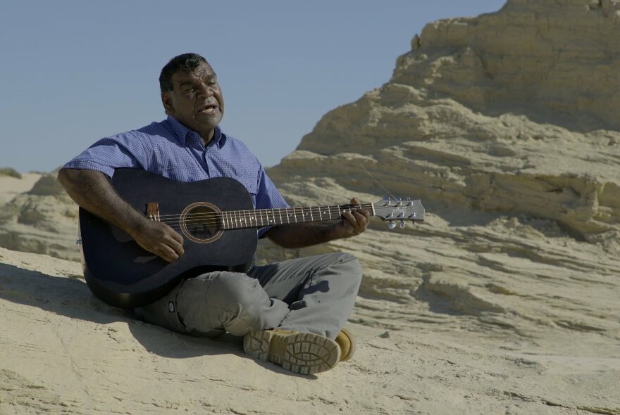 Man sitting cross legged and playing an acoustic guitar on a large sandstone rock.