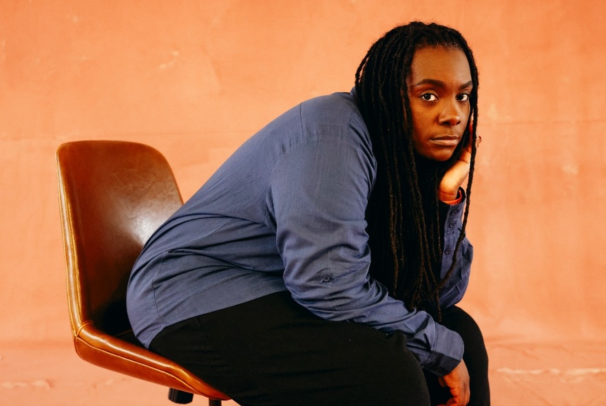 A person with long black dreadlocks, sitting in a office swivel chair and leaning forward, their upturned hand resting on their knee and supporting their chin.