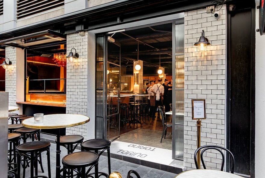 Exterior of small restaurant in a laneway, showing wide open glass doors, white brickwork, and outdoor seating, looking into a cosy and inviting interior space.