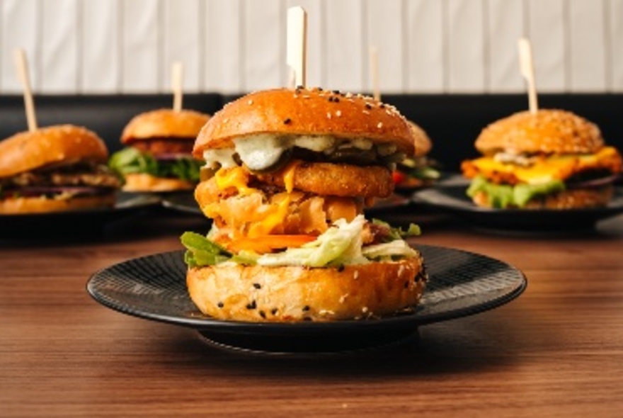 Side view of a filled jumbo burger on a plate, the bread buns held together with a skewer, with more burgers in the background.