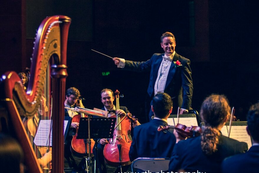 A conductor pointing his baton at a large harp while violinists and cellists play from sheet music.