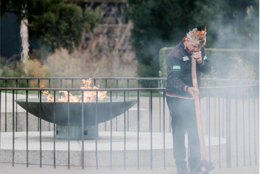 Welcome to Country ceremony at the Eternal Flame at the Shrine of Remembrance, a person wreathed in smoke standing while playing a didgeridoo.