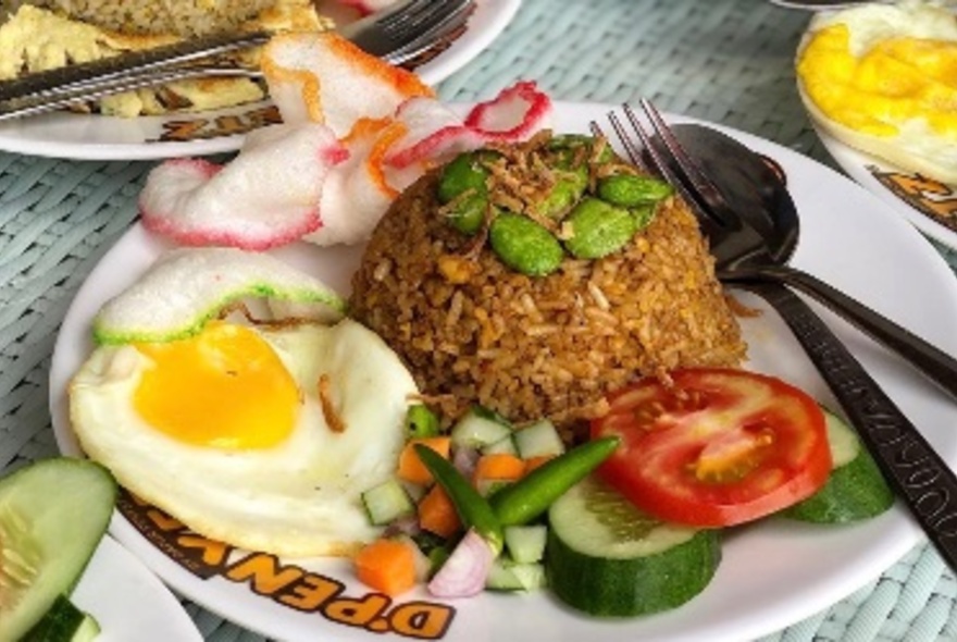 A dish of nasi goreng; a mound of fried rice, a fried egg, sliced raw tomato and cucumber, and prawn crackers. 