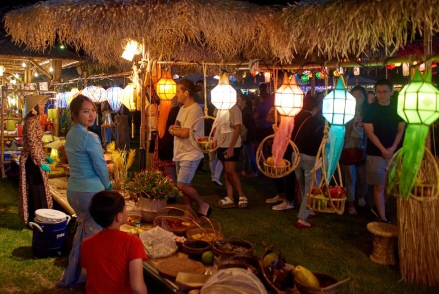 People at an outdoor street festival, some in front of food stalls, brightly coloured lanterns hanging from the stalls.