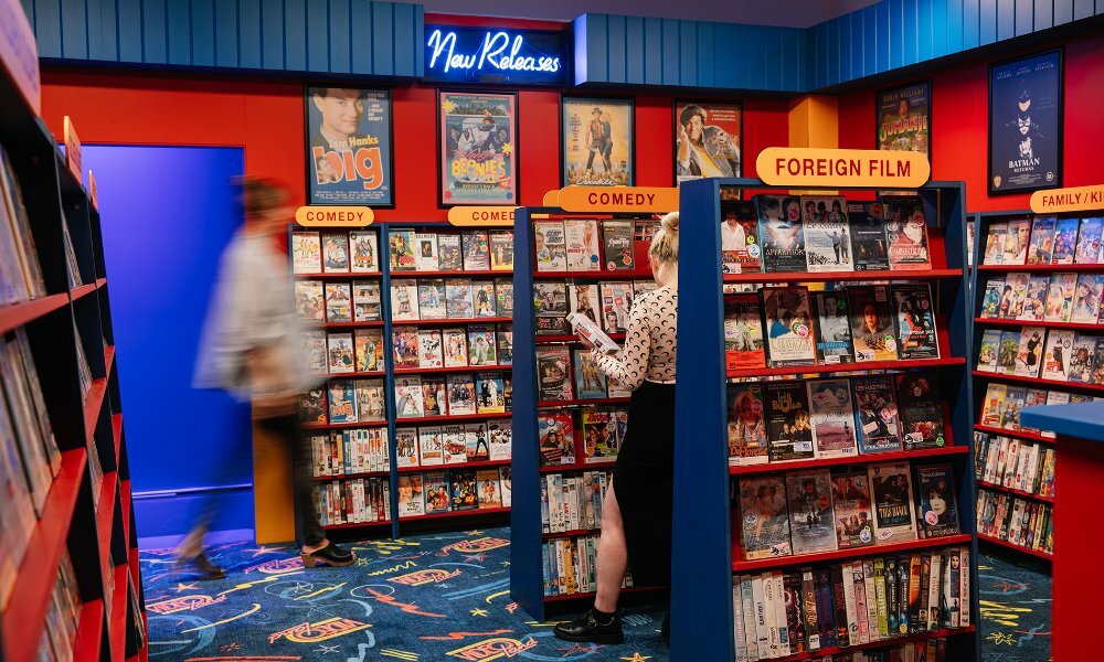 People browsing racks of video tapes in a retro video rental store art exhibition.
