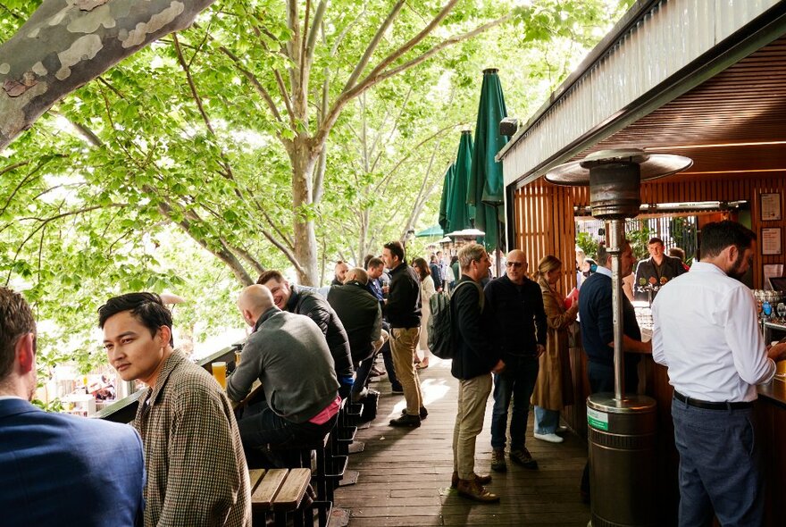 People seated and standing on the outdoor deck at Arbory Bar and Eatery, beside the Yarra River, with leafy green trees in the background.
