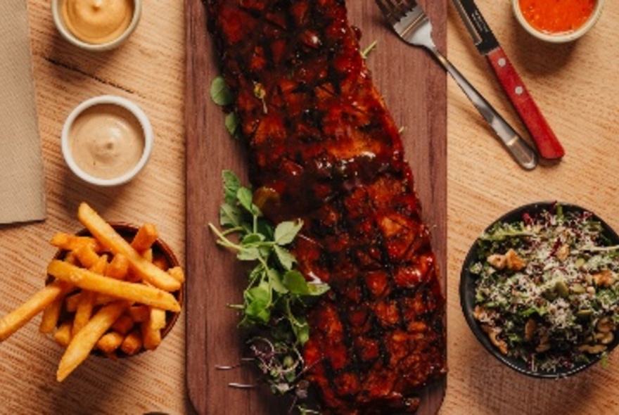 Looking down at a grilled piece of meat on a wooden tray, with fries, a salad and condiments alongside.