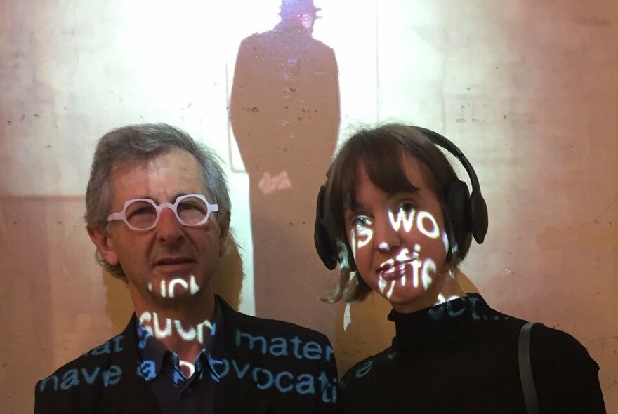 Two people in a room, with a video being screened on the wall behind them, projected over their faces. 