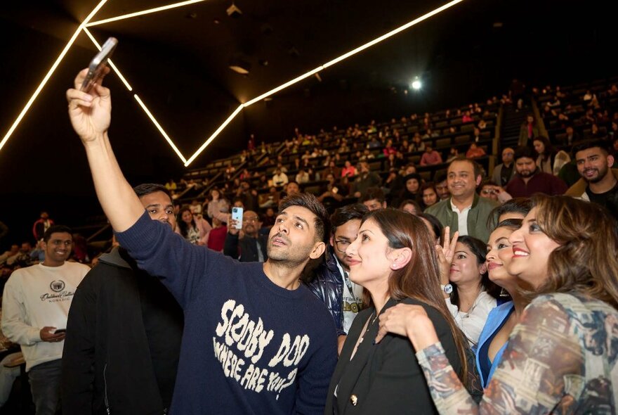 A large audience of people with a man taking a selfie of a group of women. 