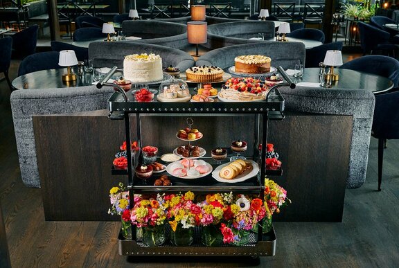 A dessert trolley with lots of cakes and flowers on it in a dark restaurant.