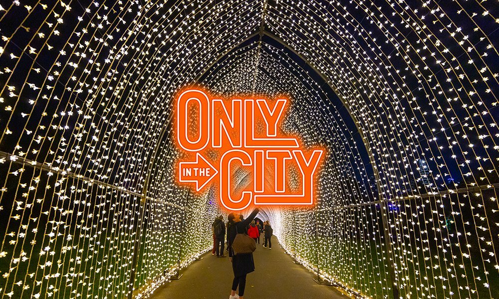 People walking through a tunnel of glowing fairy lights at night with an orange text overlay reading 'only in the city'.