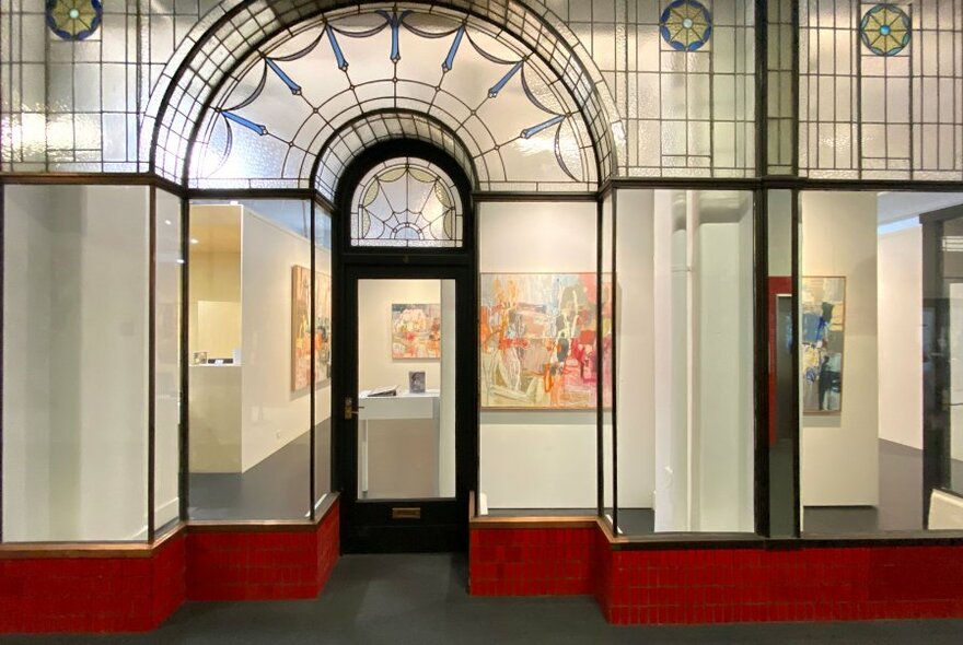 The exterior of an art gallery with stained glass arch above the door, art on the walls inside and red bricks below the windows 