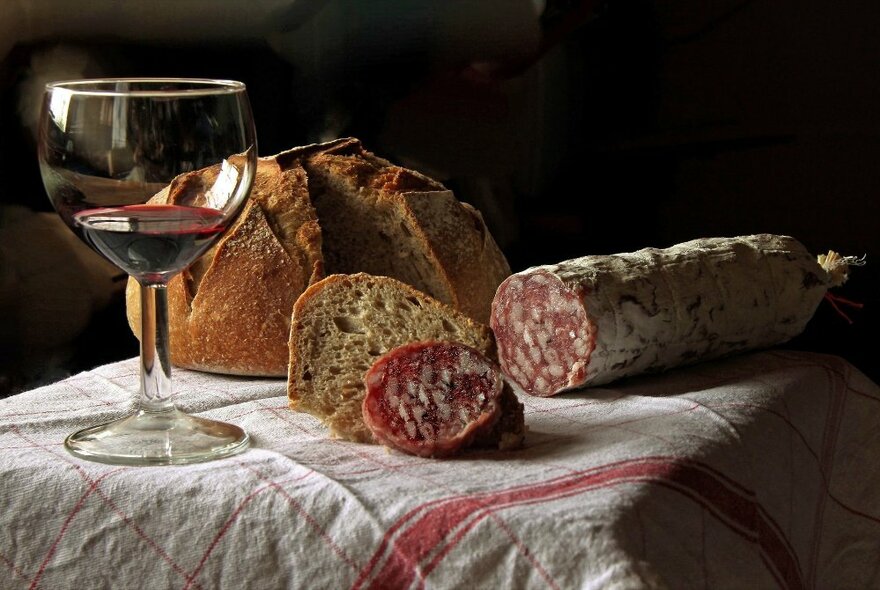 A glass of red wine on a tablecloth with bread and salami.