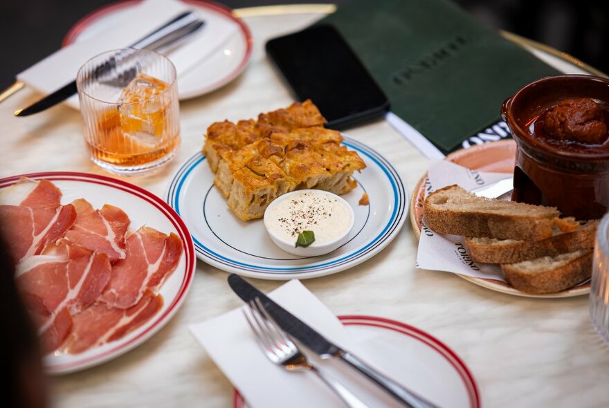 Overhead view of a table with plates of food that include prosciutto, bread and tapenade, frittata, cutlery and white napkins. 