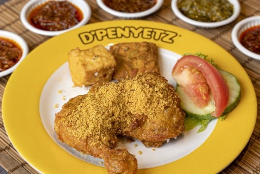 Fried chicken with tofu cubes and cucumber served on a plate that has the words D'Penyetz written on the yellow rim.