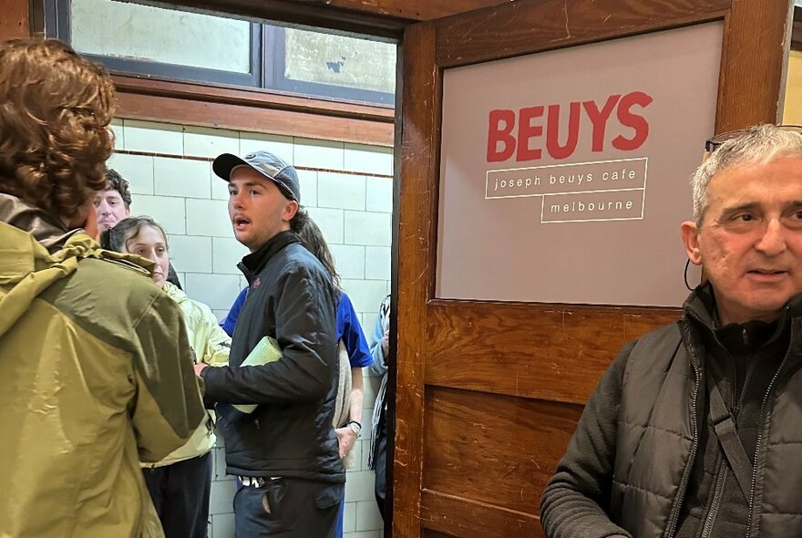 A group of people standing around in a hallway, outside a an open door into a studio, that has the word BEUYS printed on it in red letters.