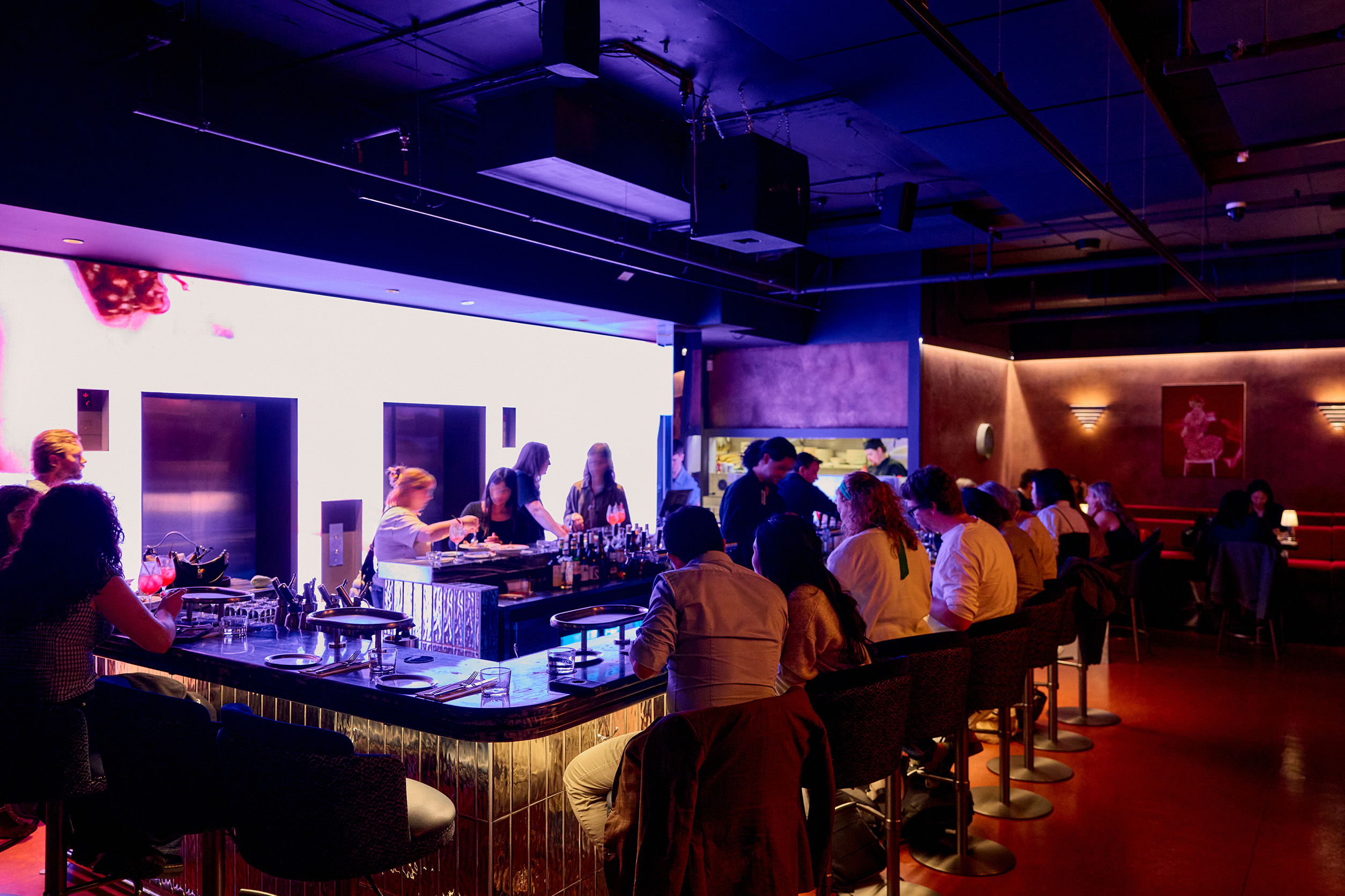 A busy restaurant with people dining and drinking at a bar surrounded by purple neon lights