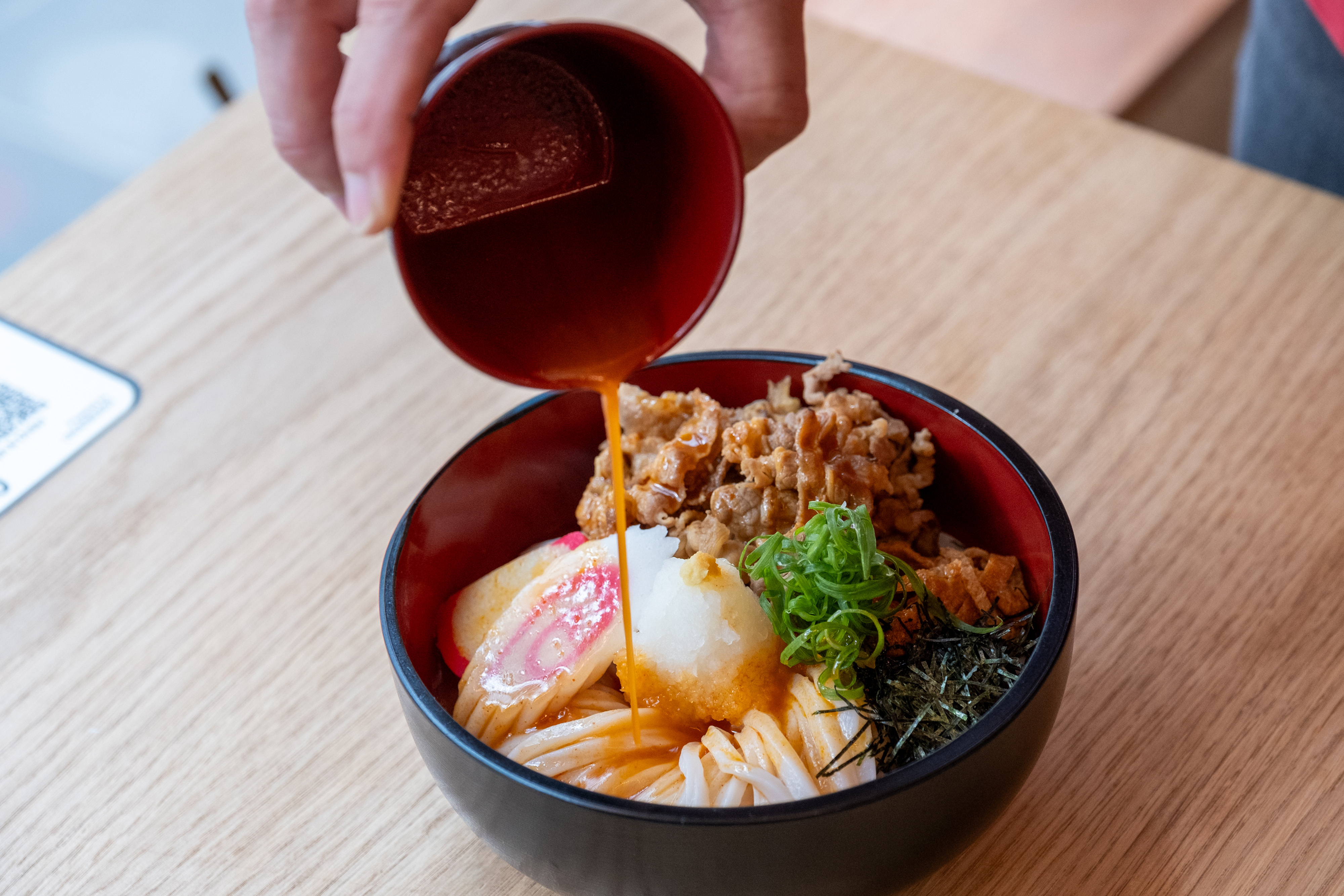 A person is pouring sauce over a bowl of udon noodles with beef, spring onions, seaweed, radish and fish cake