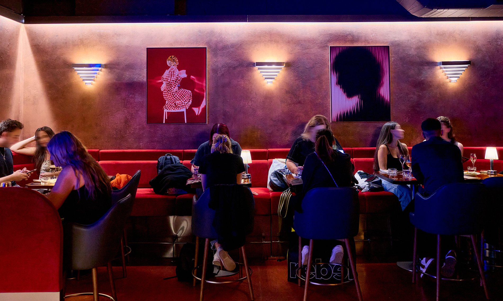 People sitting in a moody restaurant with art on the walls and long red bench seating.