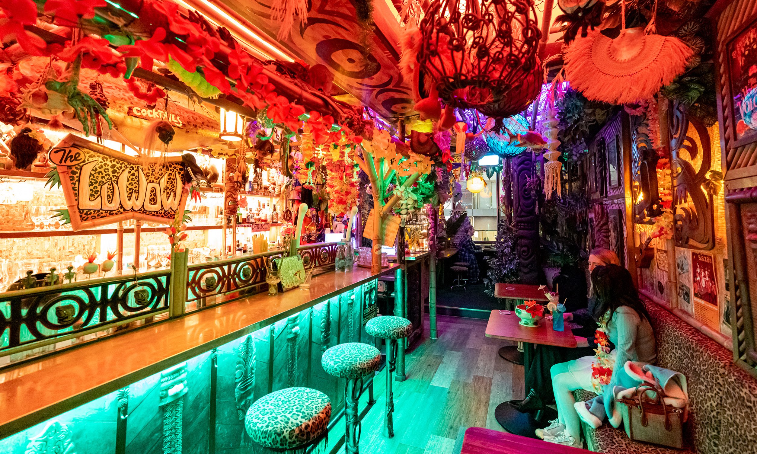 Two friends are drinking cocktails inside a kitschy tiki bar covered in tropical decorations.
