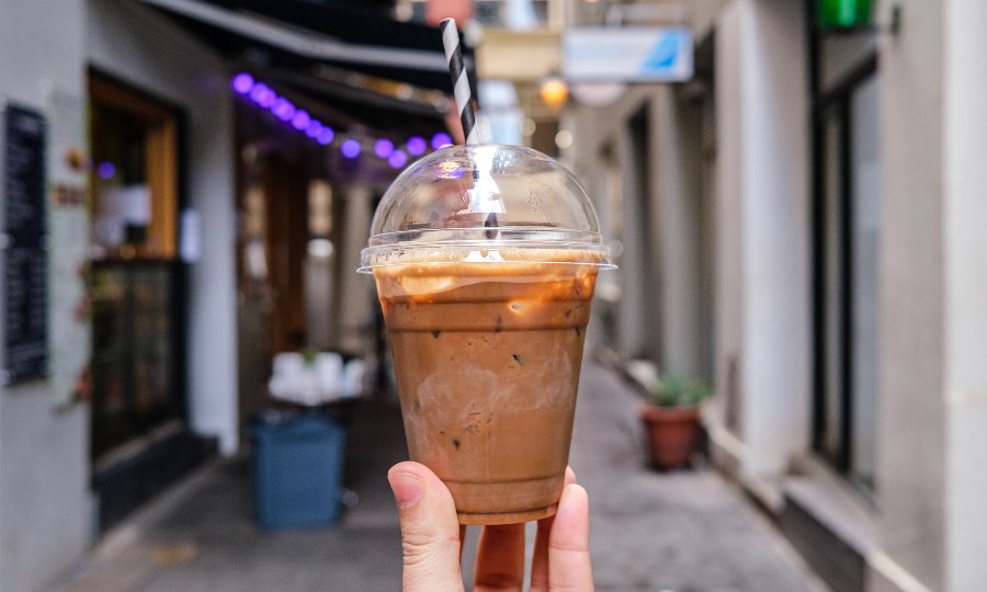 A person holding up an iced coffee with a dome lid in a laneway.
