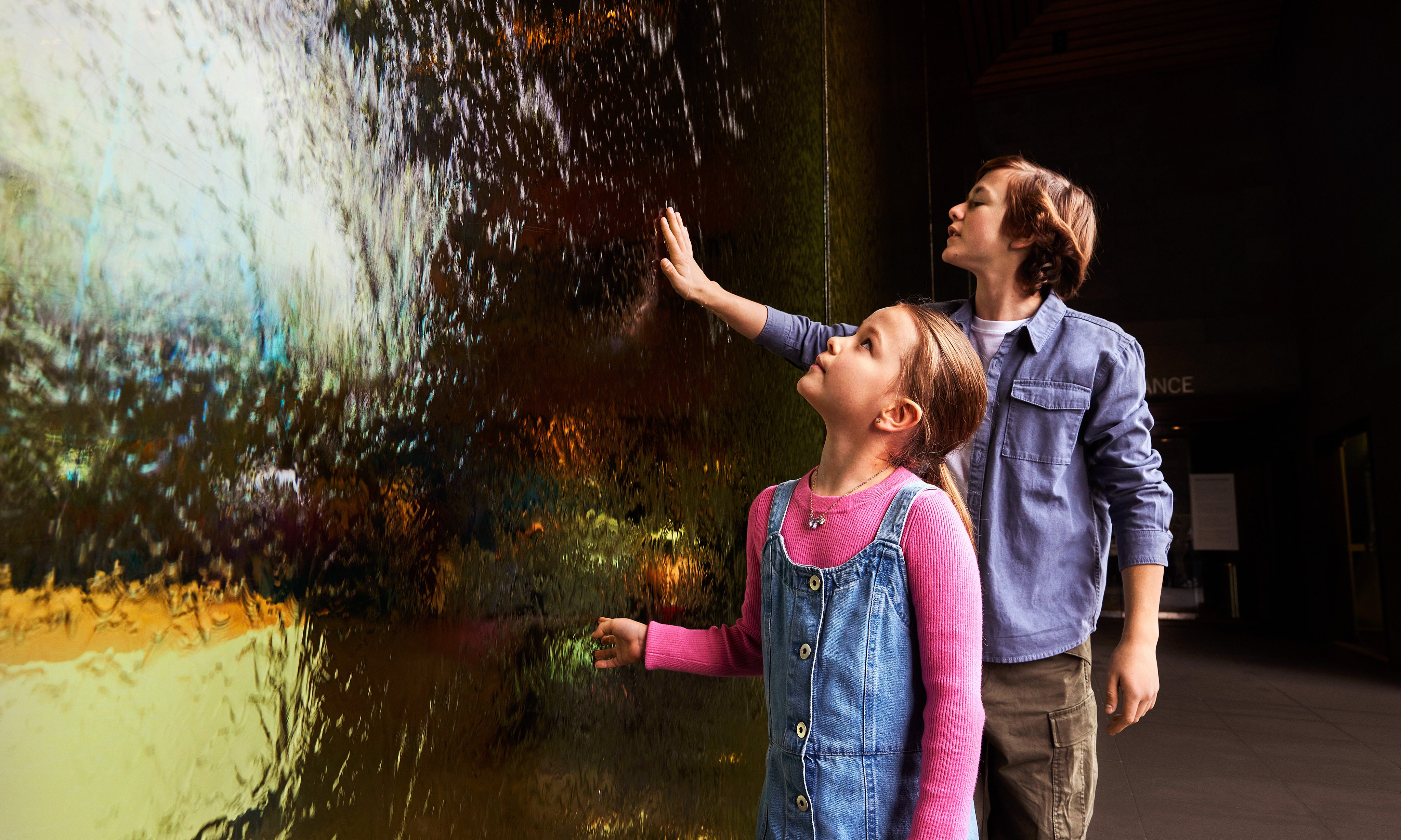 A girl and a boy reaching their hands out to touch a glass wall water feature.