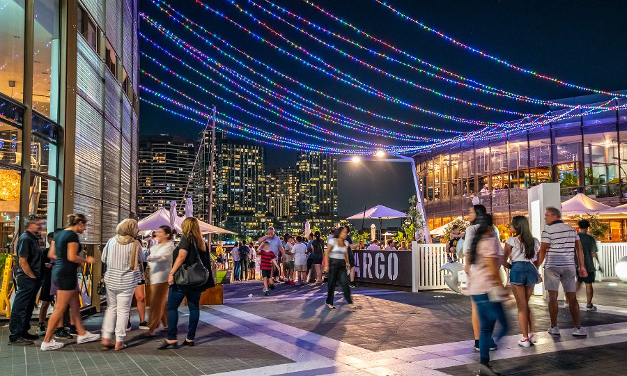 Crowds outside restaurants at night with colourful lights above them.