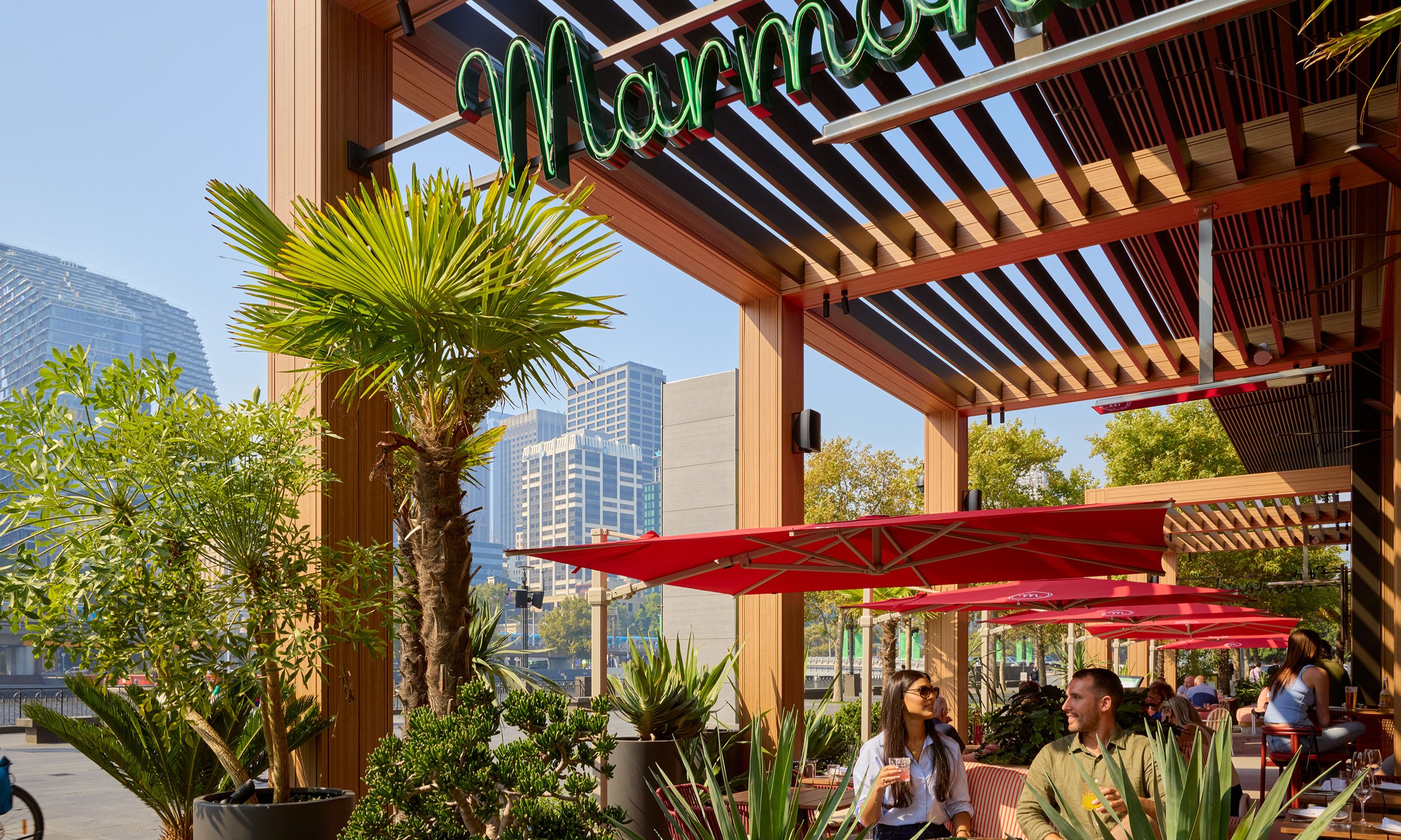 Two friends are sitting outside at a bar drinking cocktails on a sunny day. There are surrounded by pot plants.
