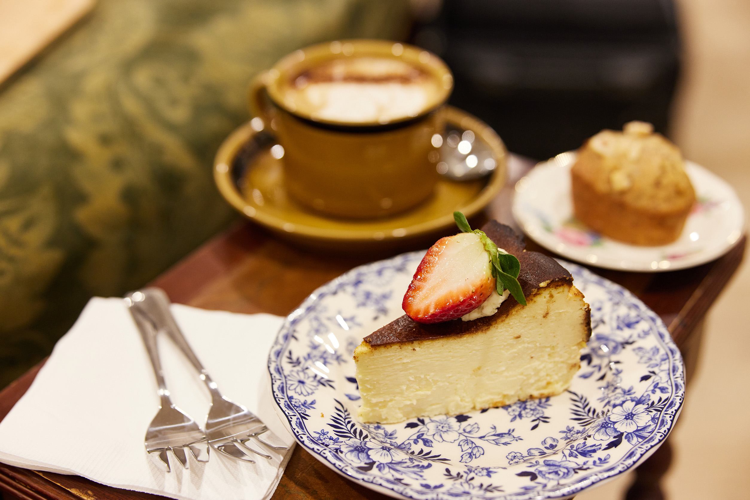 A slice of cheesecake on a decorative blue plate, behind it is a cup of coffee and a friand 