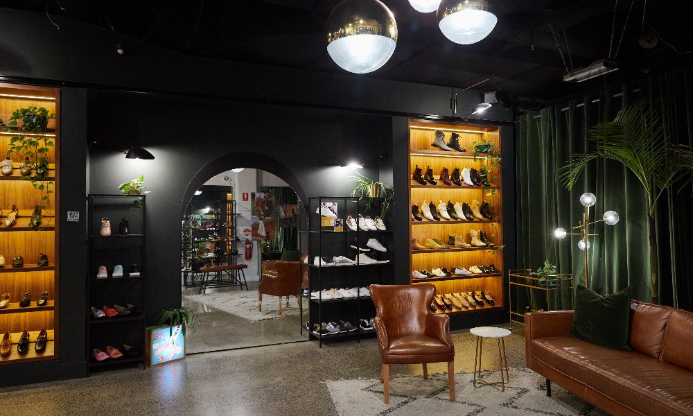 A dimly lit shoe store with black walls, illuminated shelves and tan leather furniture.