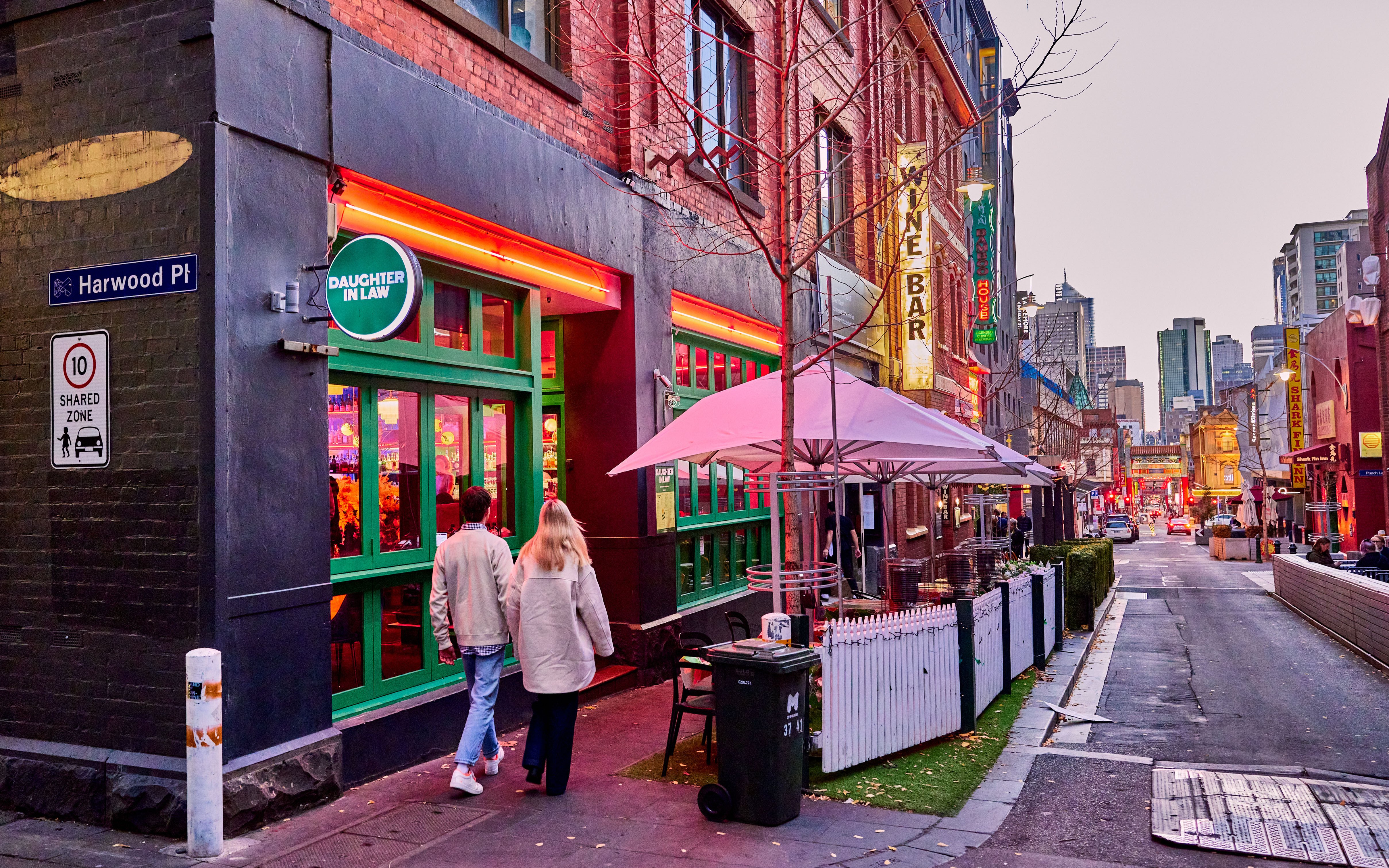 Two people walking down Chinatown outside a restaurant with green window panes and neon lighting.