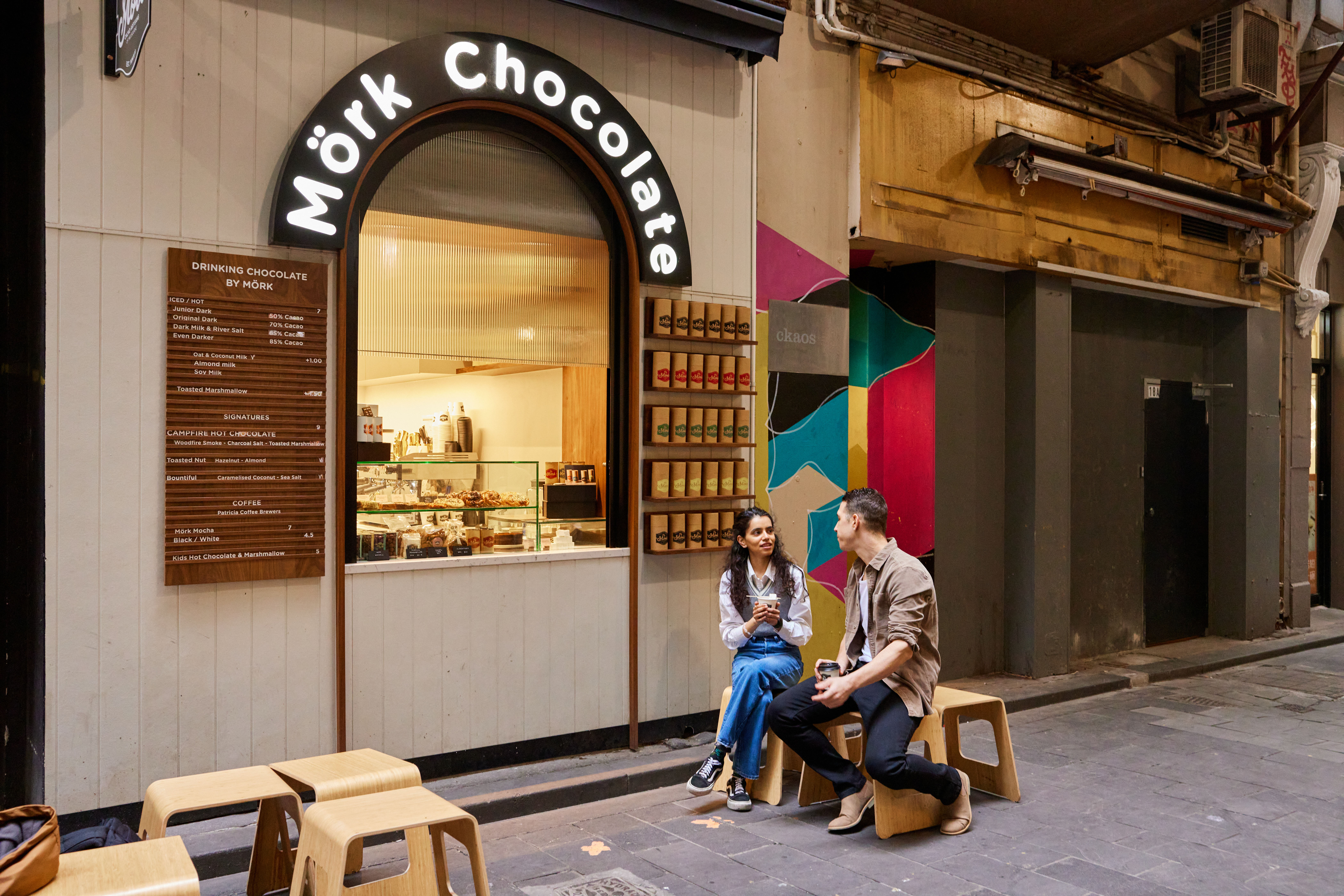 A man and woman sitting on stools outside a hole-in-the-wall cafe in a laneway.
