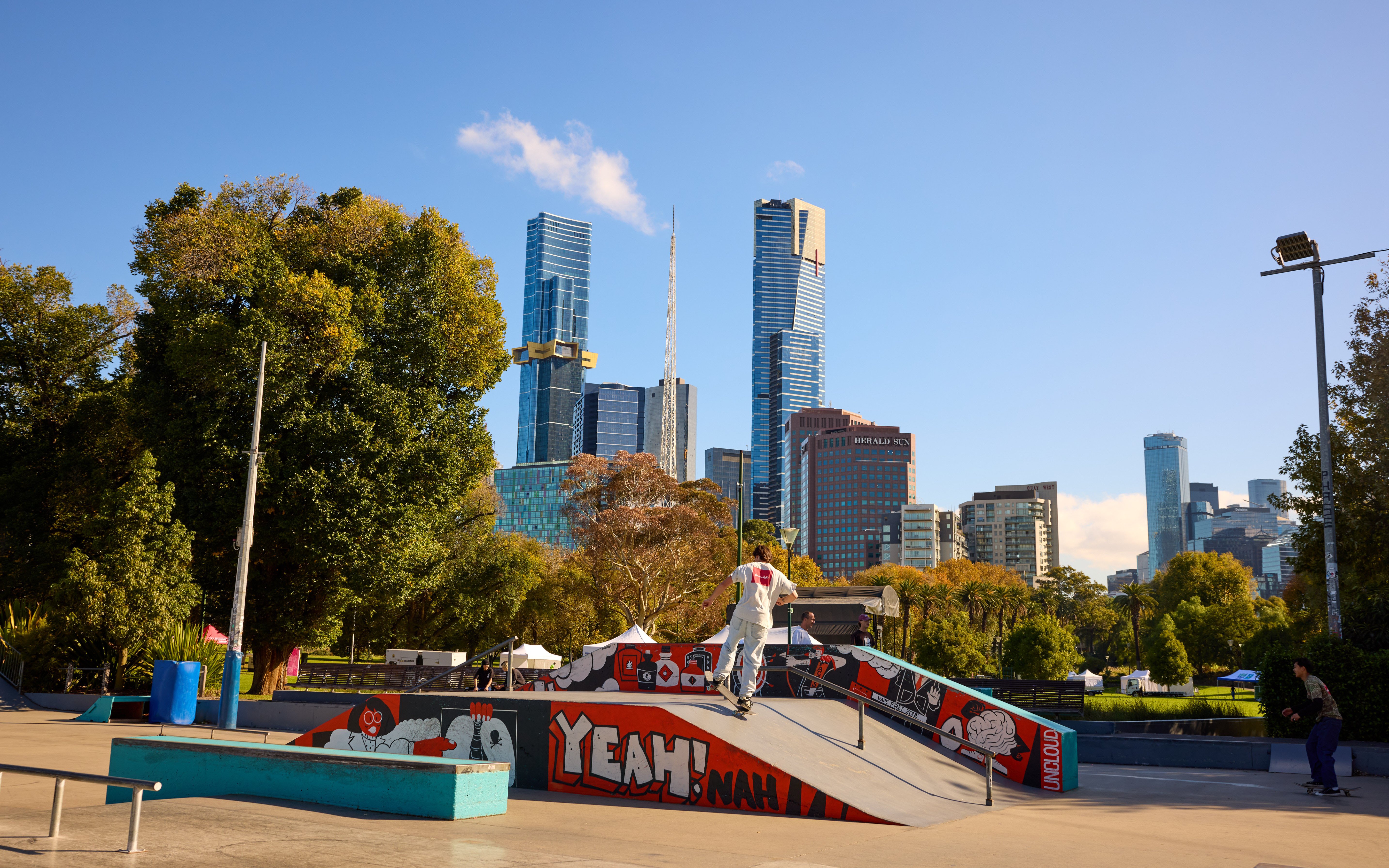 A man riding a skateboard up a ramp at a skate park with a view of the Melbourne city skyline in the background.