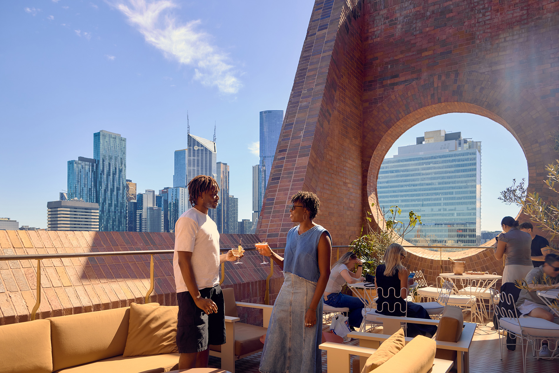 Two people drinking cocktails on an open air city rooftop bar with a brick wall with circular window.