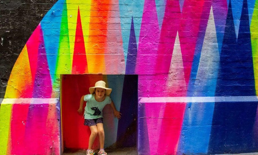 A child posing in an alcove in a rainbow mural.