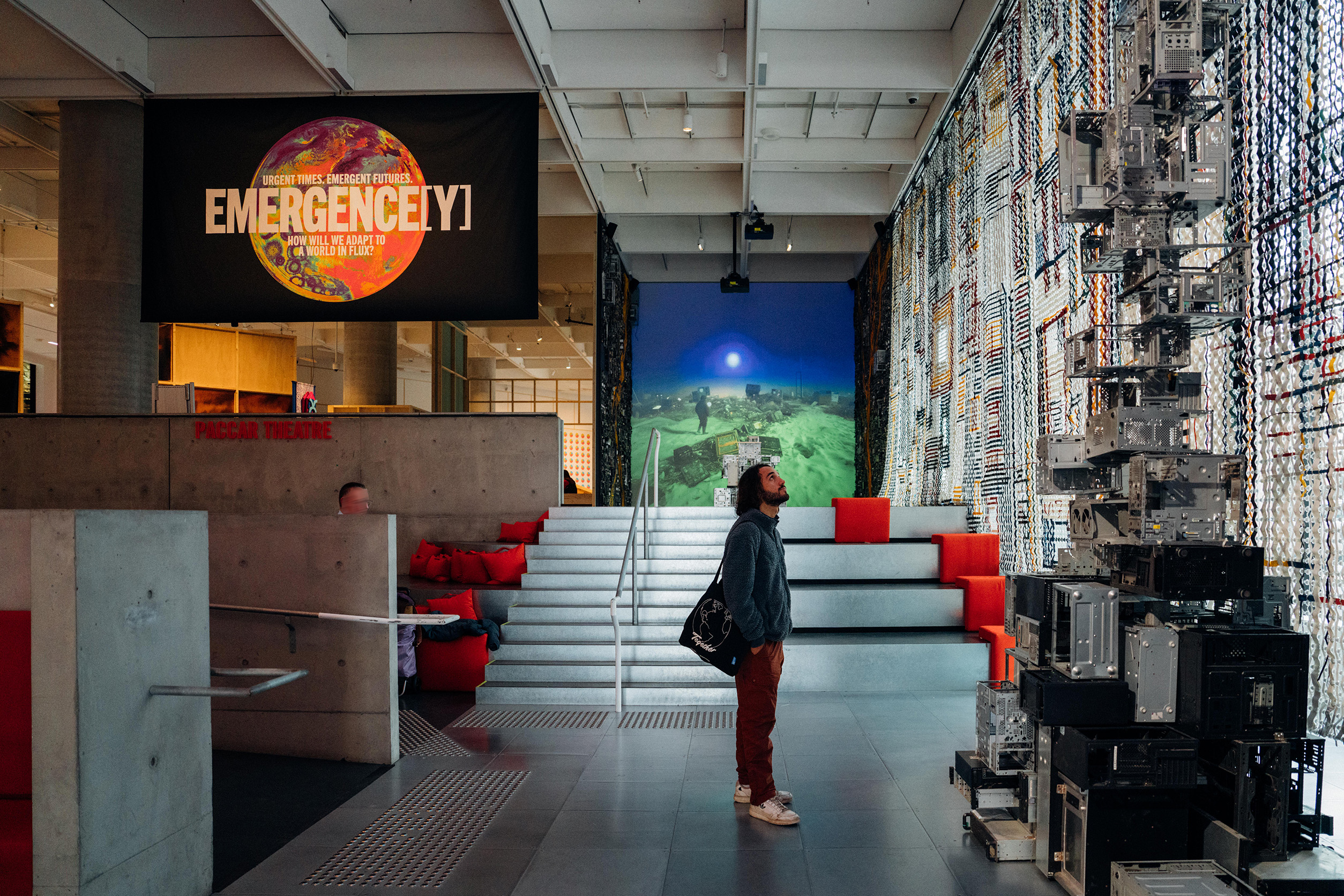 A person is inside a modern art gallery looking up at a towering sculpture behind them is a large banner saying emergency 