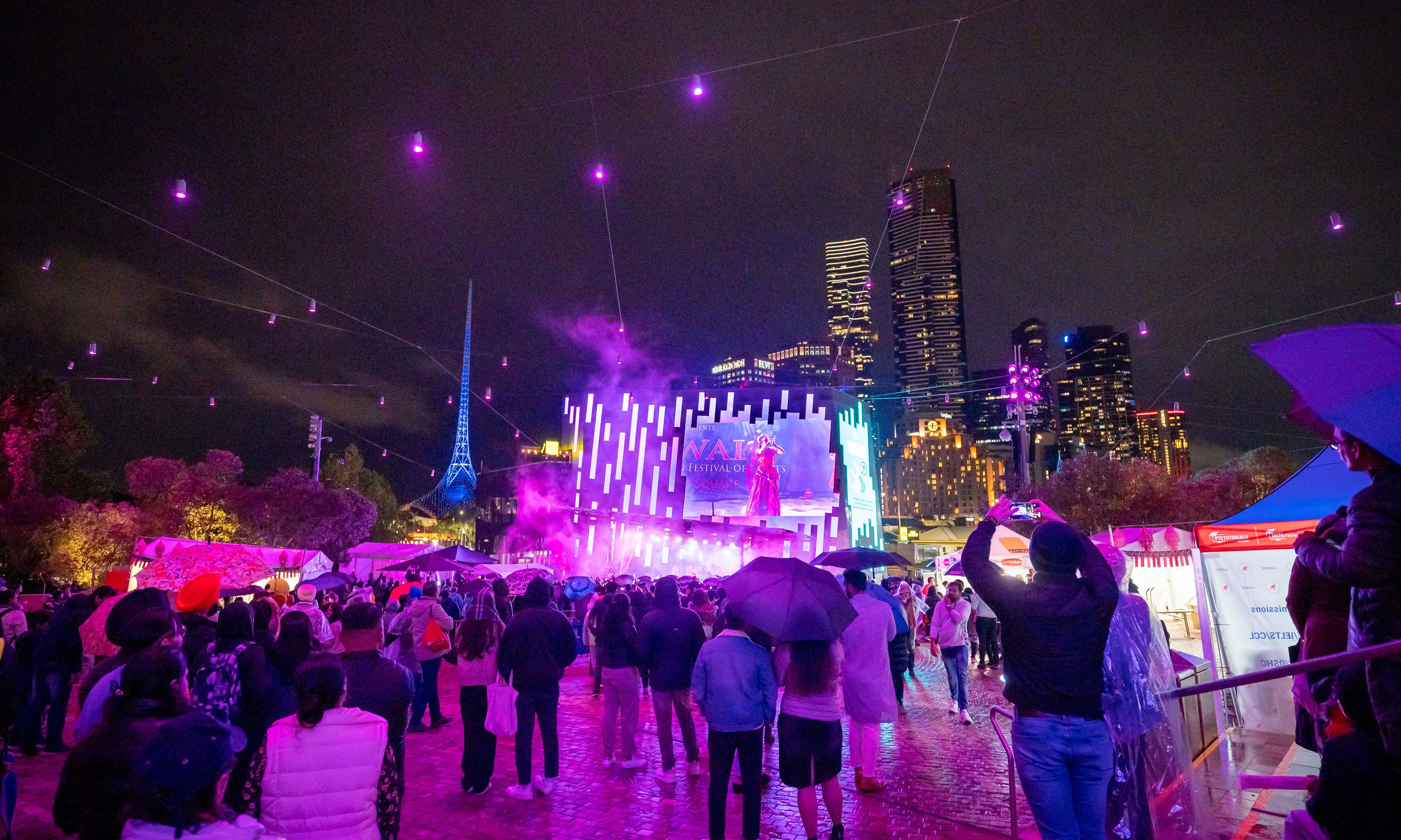 A crowd of people with umbrellas watching a purple-lit stage outdoors.