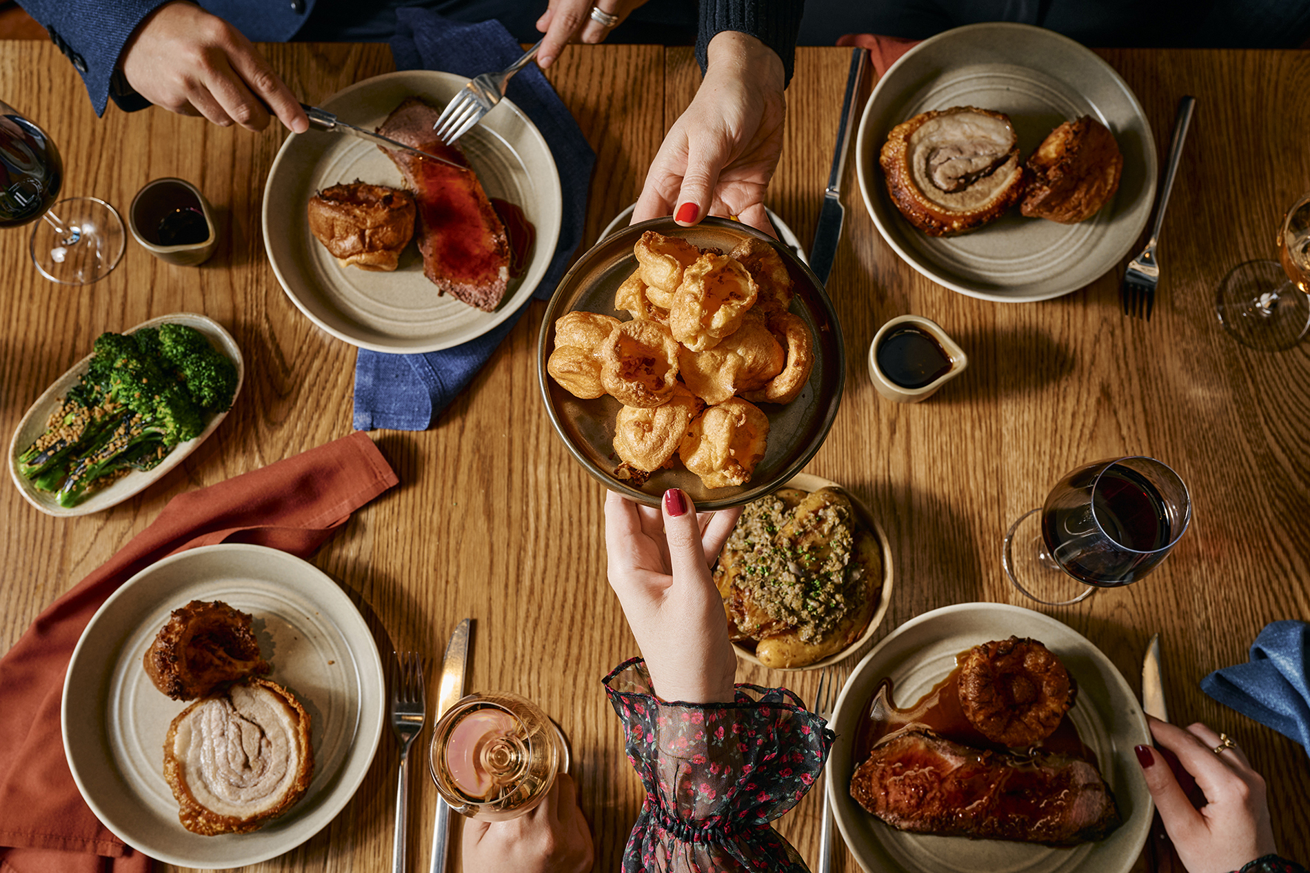 Someone passing a plate of Yorkshire puddings over a table set with rolled pork roast, veggies, wine and gravy.