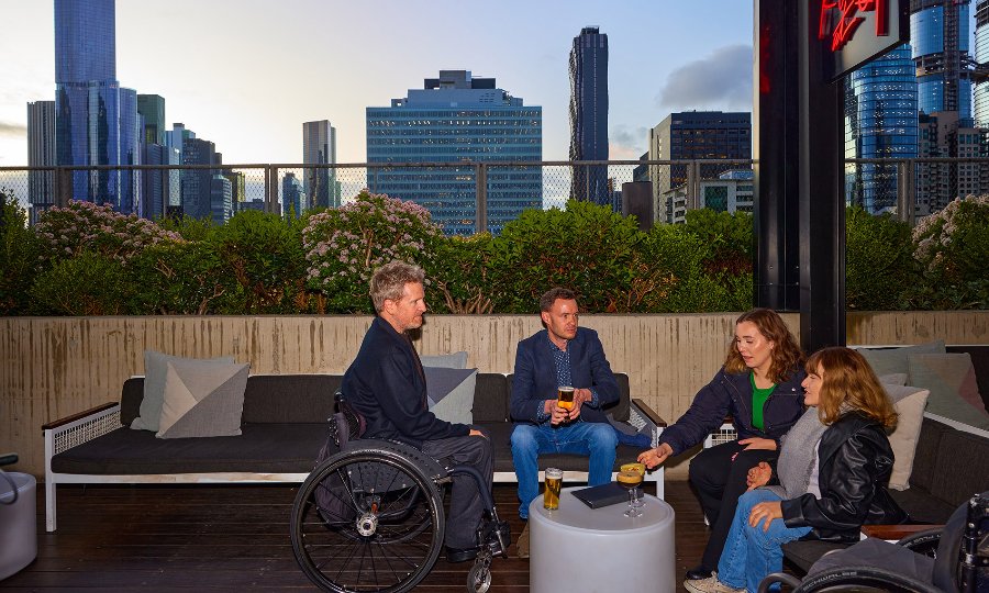 A group of friends drinking beer and cocktails on a rooftop overlooking the city skyline. Two of the group are sitting in wheelchairs.
