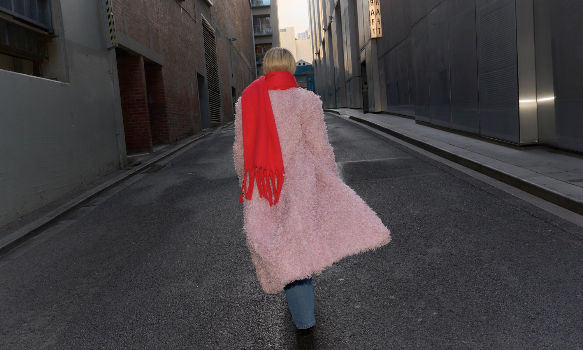 A woman is walking down an alley way wearing a pink coat and a red scarf